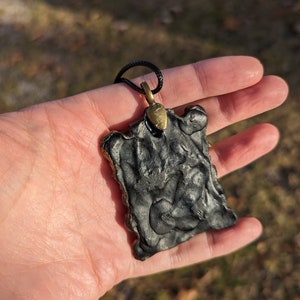 May include: Black pendant with a textured surface and a gold-colored metal bail. The pendant is hanging from a black cord.