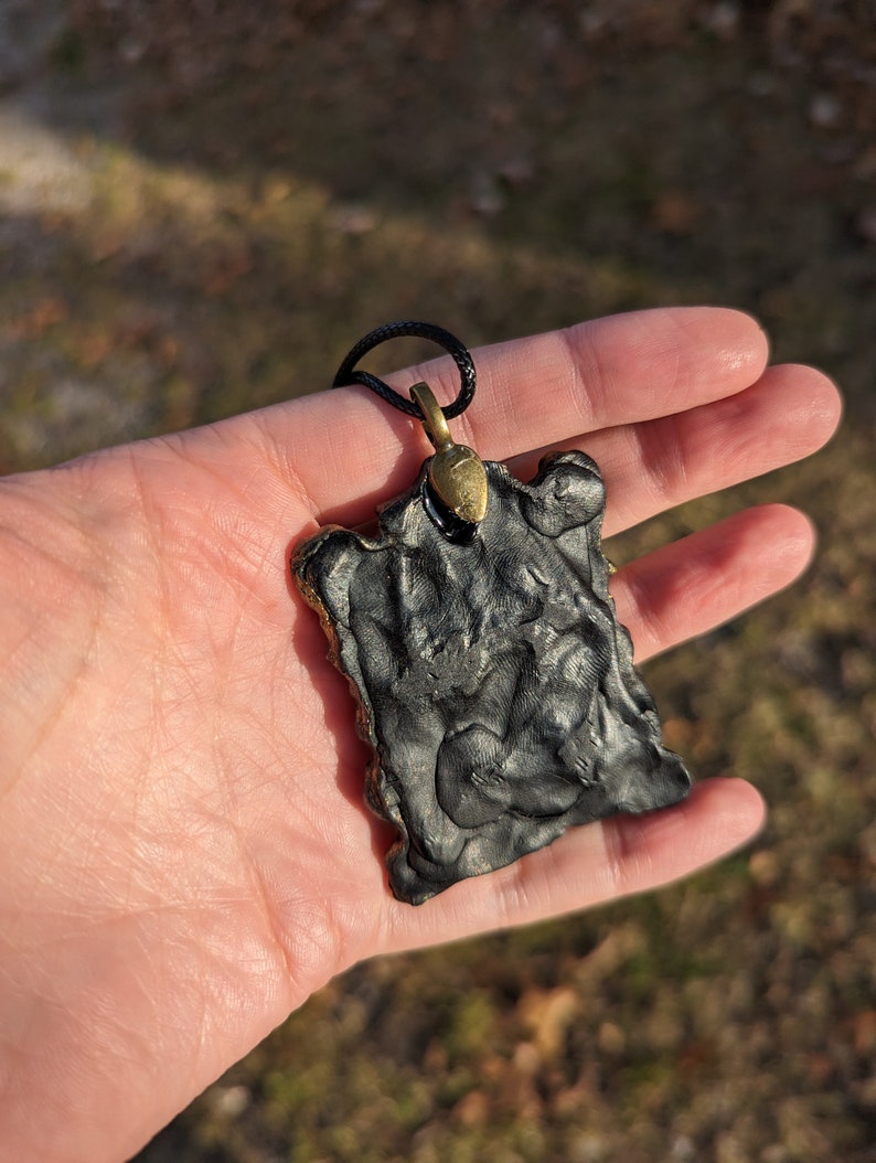 May include: Black pendant with a textured surface and a gold-colored metal bail. The pendant is hanging from a black cord.