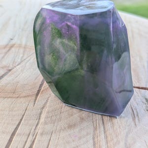 May include: A faceted, abstract resin sculpture with a mix of purple, green, and gold hues. The top edge is a lighter color, and the piece has a polished, glossy finish. The sculpture is displayed on a wooden surface.