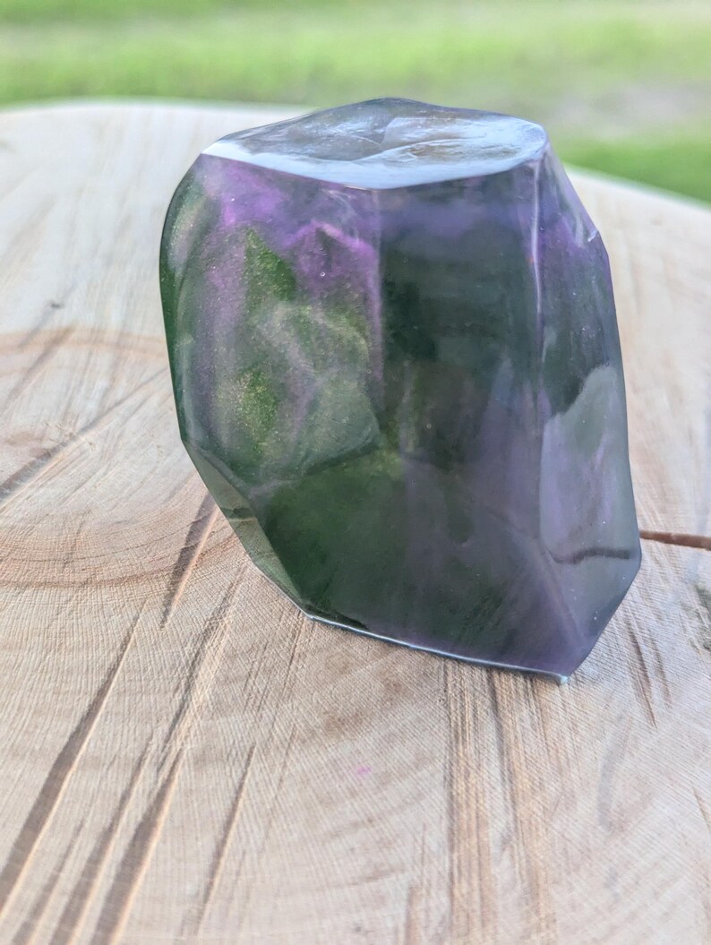 May include: A faceted, abstract resin sculpture with a mix of purple, green, and gold hues. The top edge is a lighter color, and the piece has a polished, glossy finish. The sculpture is displayed on a wooden surface.