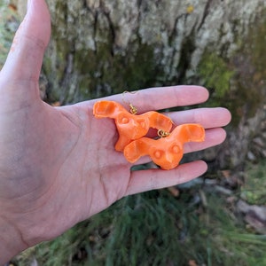 May include: A pair of bright orange earrings with a unique, abstract design. The earrings are held in a hand, with a blurred background of a tree trunk and green grass. The earrings have gold-colored hooks.