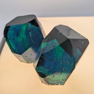 May include: Two faceted geometric sculptures made of translucent resin. The objects feature a deep black and vibrant teal blue color palette with swirling patterns and numerous small air bubbles trapped within the glossy, angular surfaces.