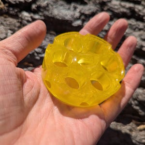 May include: A clear, yellow resin coaster with a circular design and several holes. The coaster is held in a hand, highlighting its shape and colour. The resin has a subtle shimmer, enhancing its appearance.