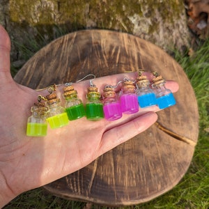 May include: A set of six colorful glass bottle earrings with cork stoppers. The bottles are filled with different colored liquids, including green, yellow, pink, and blue.