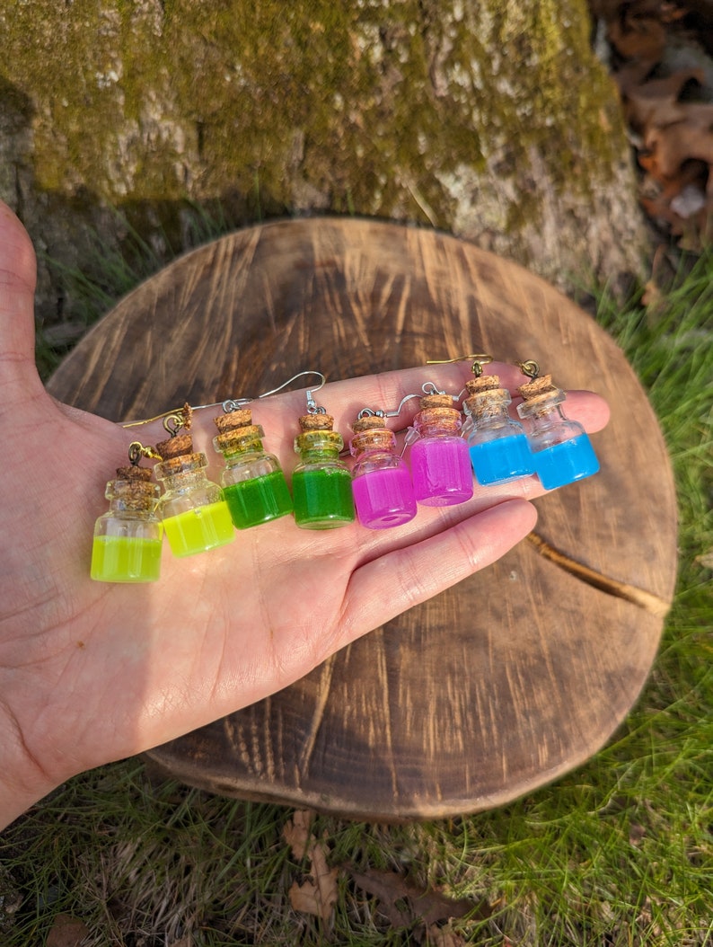 May include: A set of six colorful glass bottle earrings with cork stoppers. The bottles are filled with different colored liquids, including green, yellow, pink, and blue.
