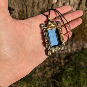 May include: A gold-colored ornate frame pendant necklace with a small mirror inside. The necklace is on a black cord.