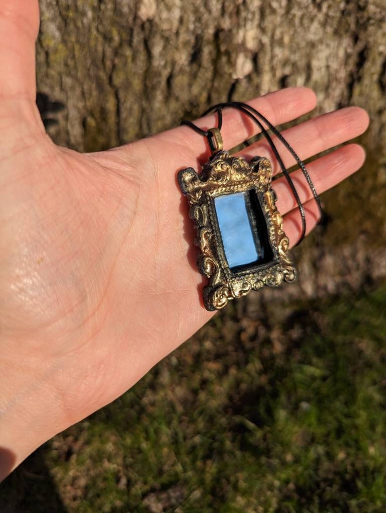 May include: A gold-colored ornate frame pendant necklace with a small mirror inside. The necklace is on a black cord.