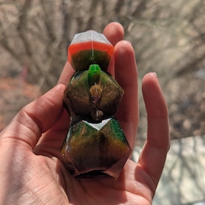 May include: A colorful, geometric resin sculpture. The sculpture is composed of stacked, faceted shapes in shades of green, brown, orange, and white. A small green accent is visible on the middle section. The sculpture is held in a hand.
