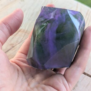 May include: A faceted, geometric-shaped object with swirling purple and green hues. The object is held in a hand, showcasing its polished, translucent appearance. The design resembles a gemstone or decorative art piece.