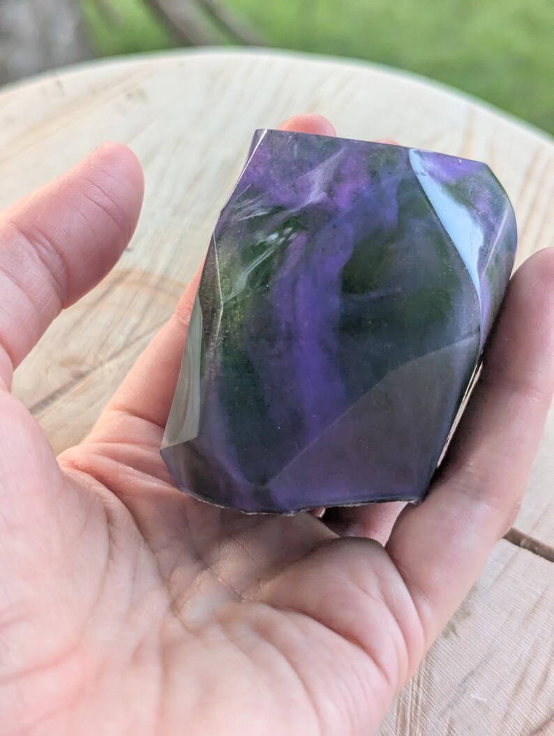 May include: A faceted, geometric-shaped object with swirling purple and green hues. The object is held in a hand, showcasing its polished, translucent appearance. The design resembles a gemstone or decorative art piece.