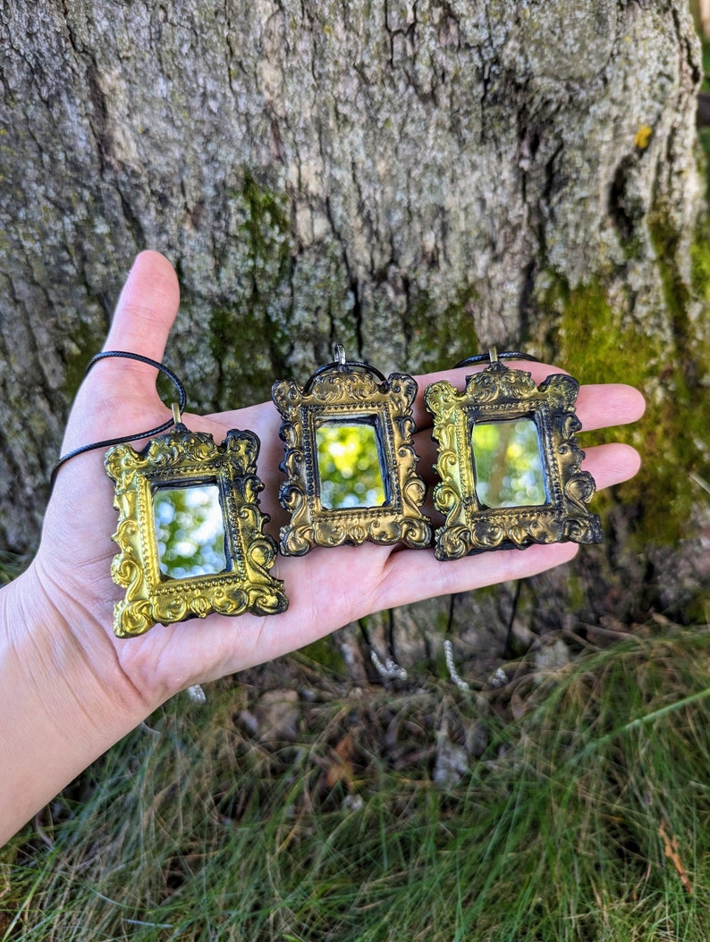 May include: Three gold-framed miniature mirrors with black accents hang from black cords. The mirrors are ornate and have a vintage aesthetic.