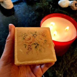 May include: A small, square, beige box with a floral design painted on the top. The box is made of alabaster and has a delicate, hand-painted design of roses and other flowers in shades of pink, yellow, and green.