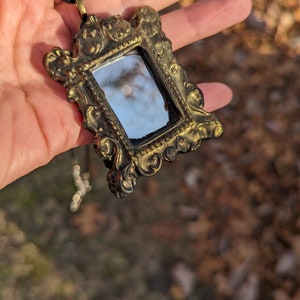 May include: A gold-toned ornate frame pendant with a small mirror inside. The frame has a detailed design with intricate carvings. The mirror reflects a blurry image of the sky.