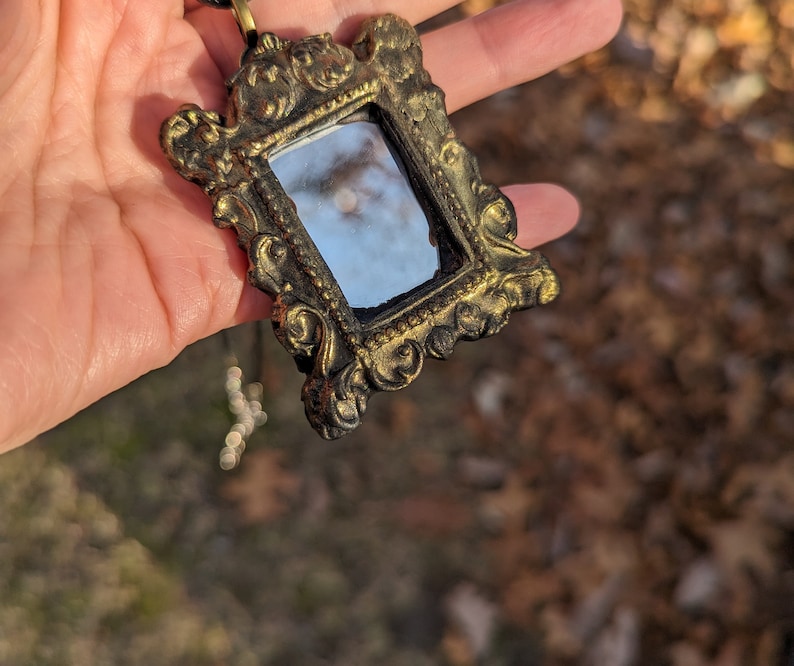May include: A gold-toned ornate frame pendant with a small mirror inside. The frame has a detailed design with intricate carvings. The mirror reflects a blurry image of the sky.