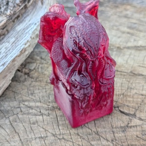 May include: A translucent, ruby-red resin sculpture of a creature with a textured head and tentacles. The sculpture is mounted on a rectangular base. The artwork is displayed on a wooden surface.