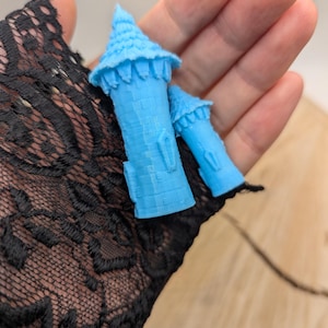 May include: Two small, light blue, 3D printed towers with detailed designs. The towers are held in a hand.