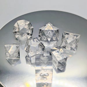 May include: A set of seven clear, faceted polyhedral dice. The dice are transparent with white numbering and are arranged on a reflective surface. The set includes a d4, d6, d8, d10, d12, d20, and a percentile die.