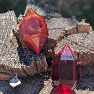 Puede incluir: Dos objetos facetados en forma de cristal hechos de resina roja translúcida. El objeto más grande presenta remolinos de purpurina dorada en su interior, mientras que el más pequeño es de un rojo profundo y claro. Diseño geométrico puntiagudo.