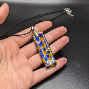 May include: A rectangular pendant necklace with a colorful mosaic design. The pendant features a mix of blue, yellow, and orange glass pieces. It hangs from a black cord with a silver-toned clasp. The pendant is approximately 1.5 inches long.