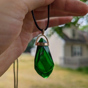 Puede incluir: Un collar con un colgante de piedra preciosa verde en forma de lágrima con un engaste de oro. El colgante está facetado. El collar está en un cordón negro.