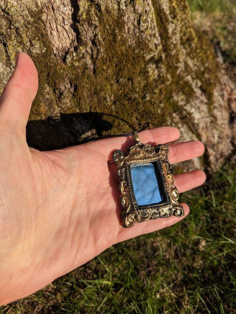 May include: A gold-toned ornate frame pendant necklace with a small mirror inside. The mirror reflects the sky and trees.