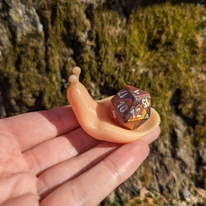 May include: A small, beige, plastic snail figurine with a brown and gold iridescent dice on its back. The dice has numbers 6, 8, 10, and 12 visible.