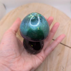 May include: A green and purple resin mushroom with iridescent glitter. The mushroom has a rounded top and a smaller, round base.