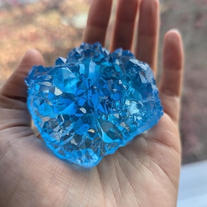 May include: A cluster of bright blue crystals, held in an open hand. The crystals have a faceted, irregular shape, catching the light and creating a sparkling effect. The overall appearance suggests a geode or mineral specimen.