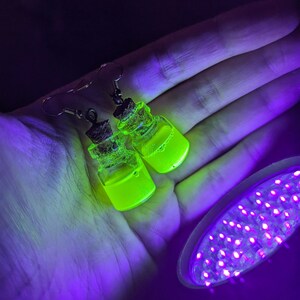 May include: A pair of earrings featuring small glass bottles filled with a bright green liquid. The bottles are attached to silver ear wires and have a cork stopper. The earrings are glowing under a black light.