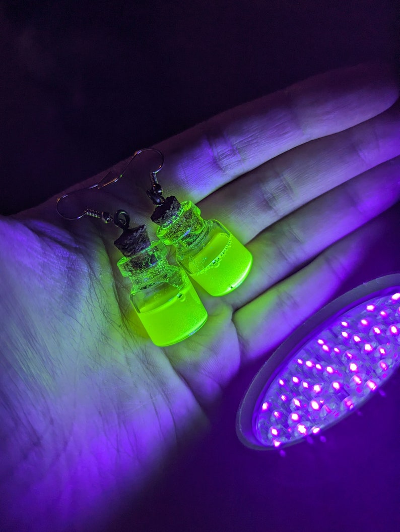 May include: A pair of earrings featuring small glass bottles filled with a bright green liquid. The bottles are attached to silver ear wires and have a cork stopper. The earrings are glowing under a black light.