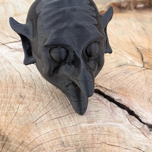Puede incluir: Una escultura negra impresa en 3D de una cabeza de duende con orejas puntiagudas y una nariz larga y puntiaguda. La escultura tiene una superficie texturizada y está colocada sobre un fondo de madera natural.