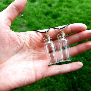 May include: Two small glass bottles with cork stoppers on black cords. The bottles are hanging from a hand.