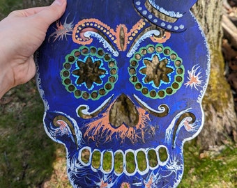 May include: A blue and white sugar skull with a black top hat. The skull has green and orange details and is painted with a marbled effect.