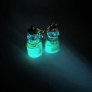 May include: A pair of earrings with small glass bottles filled with a glowing green substance. The bottles have cork stoppers and are attached to gold-colored hooks.