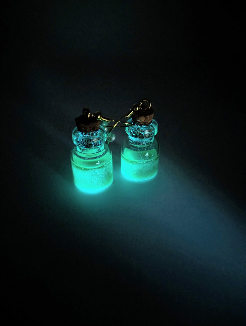 May include: A pair of earrings with small glass bottles filled with a glowing green substance. The bottles have cork stoppers and are attached to gold-colored hooks.