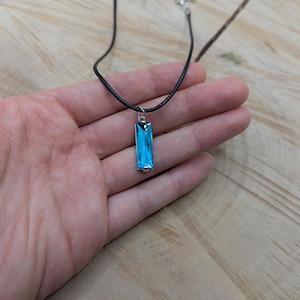 May include: A rectangular, light blue pendant necklace with a silver-colored bail. The pendant is suspended from a black cord necklace with a silver-colored clasp. The necklace is displayed on a wooden surface.