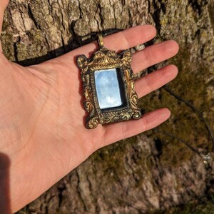 May include: A gold-colored ornate mirror pendant necklace with a black cord. The mirror is rectangular and has a detailed frame.