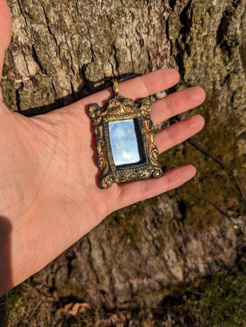 May include: A gold-colored ornate mirror pendant necklace with a black cord. The mirror is rectangular and has a detailed frame.