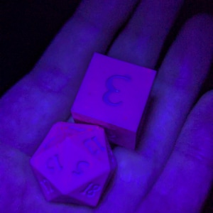 Puede incluir: Dos dados rosas, uno cúbico con el número 3 y otro de 20 caras con números visibles. Los dados se sostienen en una mano, iluminados por una luz púrpura.