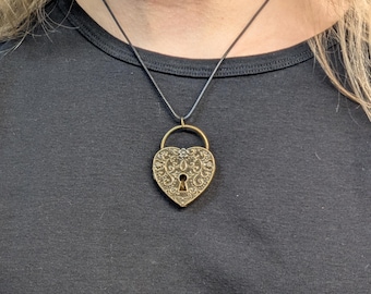 May include: A gold heart-shaped locket necklace with an intricate design and a keyhole. The necklace is on a black cord.