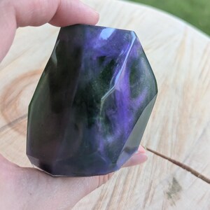 May include: A faceted, geometric-shaped decorative object with a swirling mix of purple, green, and black colors. The object is held in a hand, and the background is a wooden surface.