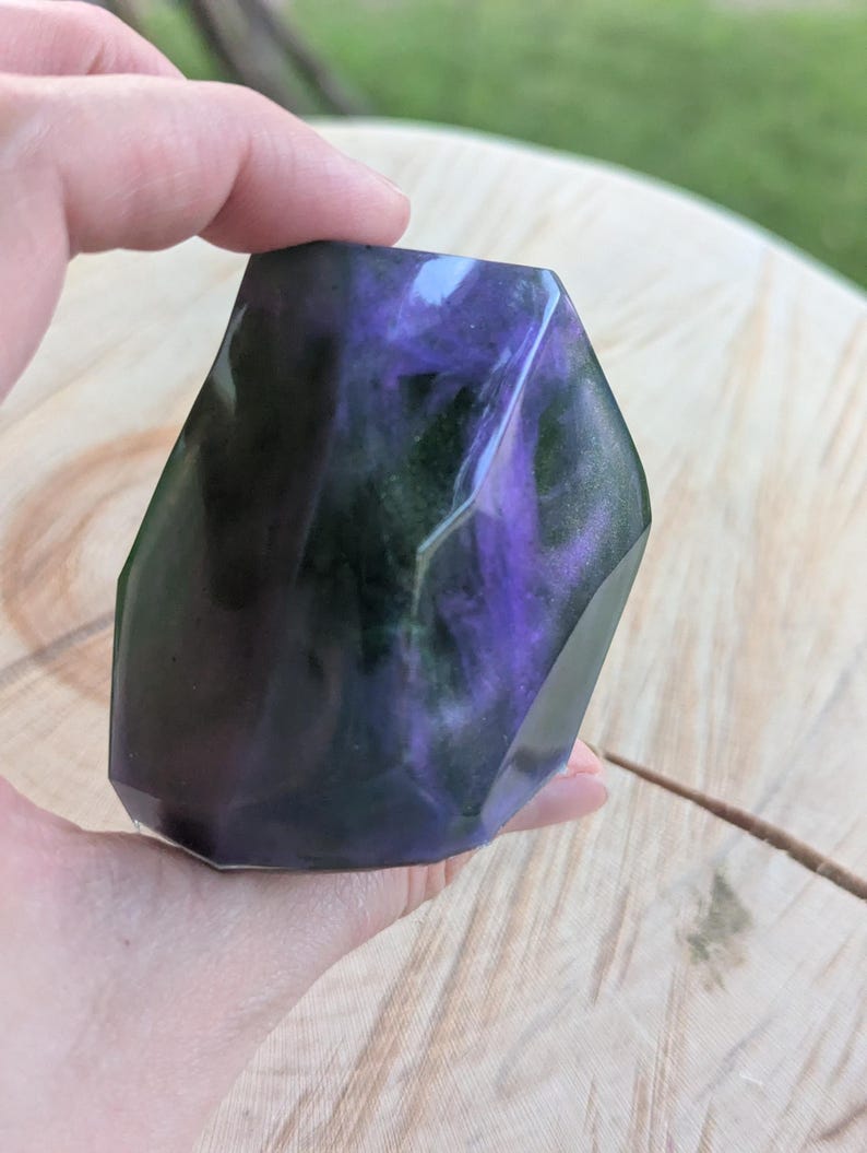 May include: A faceted, geometric-shaped decorative object with a swirling mix of purple, green, and black colors. The object is held in a hand, and the background is a wooden surface.