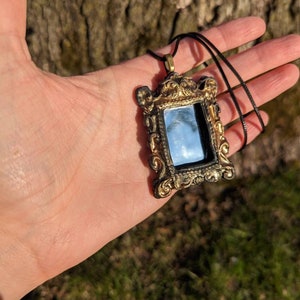 May include: A gold-colored ornate frame pendant necklace with a small mirror inside. The frame is black and gold and has a detailed design. The necklace is on a black cord.