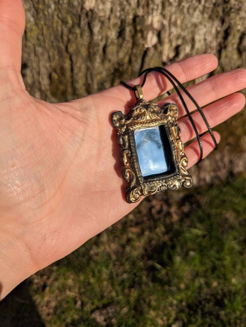 May include: A gold-colored ornate frame pendant necklace with a small mirror inside. The frame is black and gold and has a detailed design. The necklace is on a black cord.