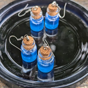 May include: Three blue glass bottle earrings with cork stoppers and silver hooks. The earrings are on a black plate.
