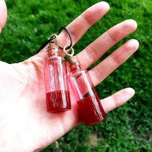 Puede incluir: Dos pequeñas botellas de vidrio transparente llenas de un líquido rojo, suspendidas de un cordón negro. Las botellas tienen tapones de corcho y aros metálicos dorados. El fondo es hierba verde borrosa.