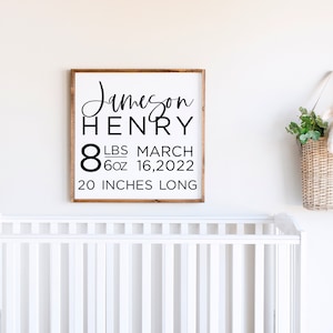 May include: A framed wooden sign with the name "Jameson Henry" in black script. The sign includes the baby's birth details: 8 lbs 6 oz, March 16, 2022, and 20 inches long. A wicker basket with a stuffed lamb and greenery hangs on the wall.