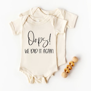 Könnte beinhalten: Zwei cremefarbene Baby-Bodys mit kurzen Ärmeln. Der obere Body hat den Text "Oops! We did it again" in Schwarz.