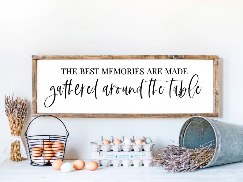 May include: A wooden sign with the text "The best memories are made gathered around the table". The sign is hanging on a white wall. There are eggs, a basket, and dried lavender in the foreground.