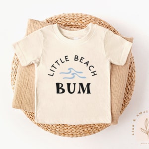以下が含まれることがあります： 黒と青の文字で「Little Beach Bum」と書かれた白い半袖Tシャツ。テキストは青い波のグラフィックで囲まれています。